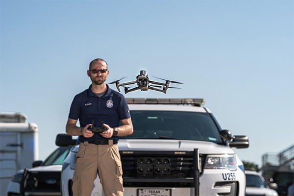 Policjant stojący obok wozu policyjnego, nad którym unosi się Dron DJI Mavic 3 Thermal Advanced C1