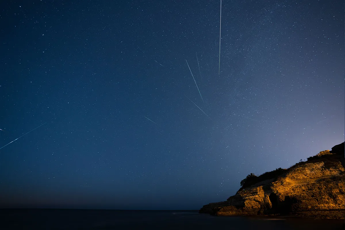 Jasne ślady meteorów na nocnym niebie nad skalistym wybrzeżem.